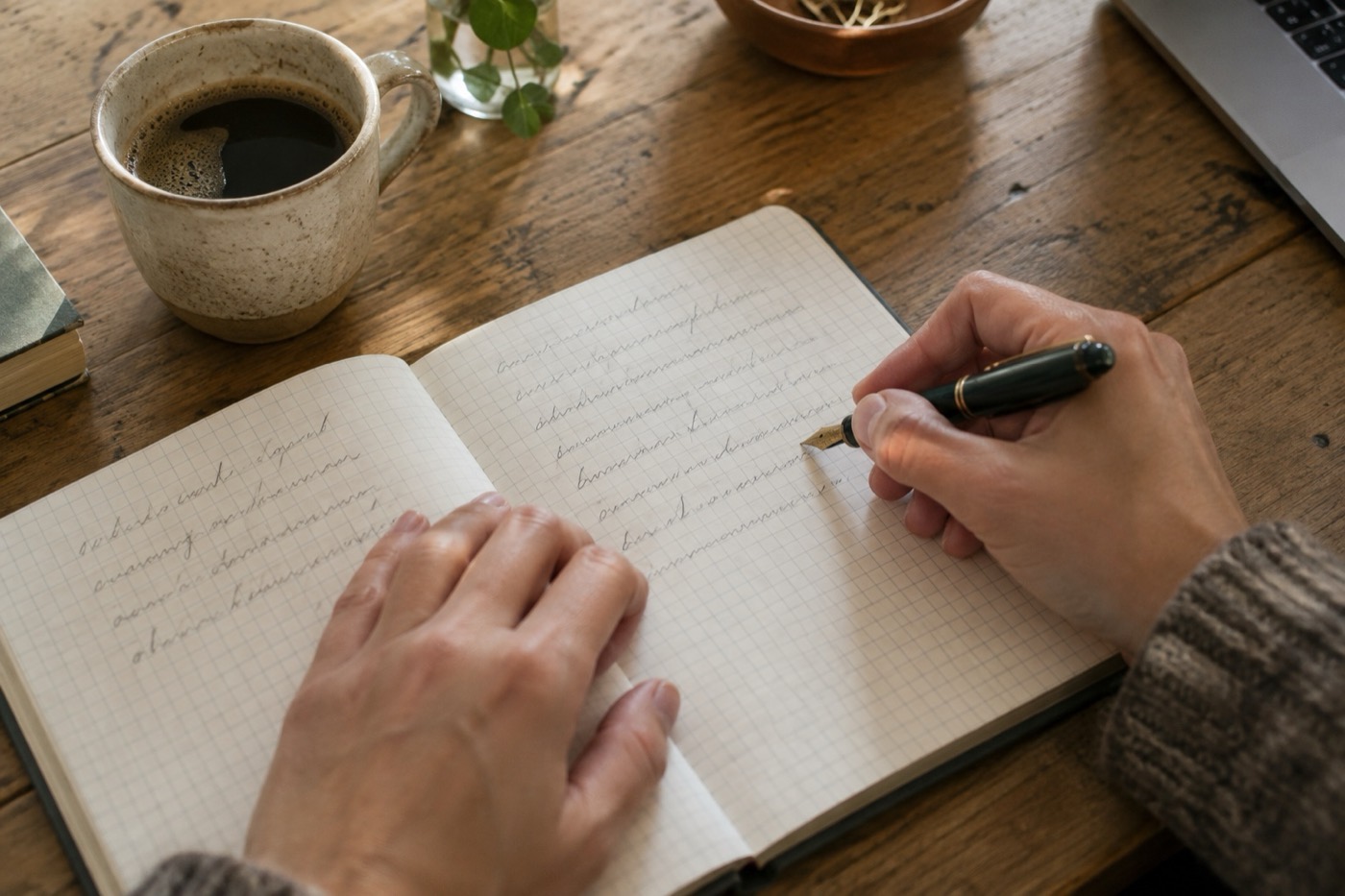 木の机の上で、開いたノートに万年筆で文章を書いている手元。横にはコーヒーのカップが置かれている